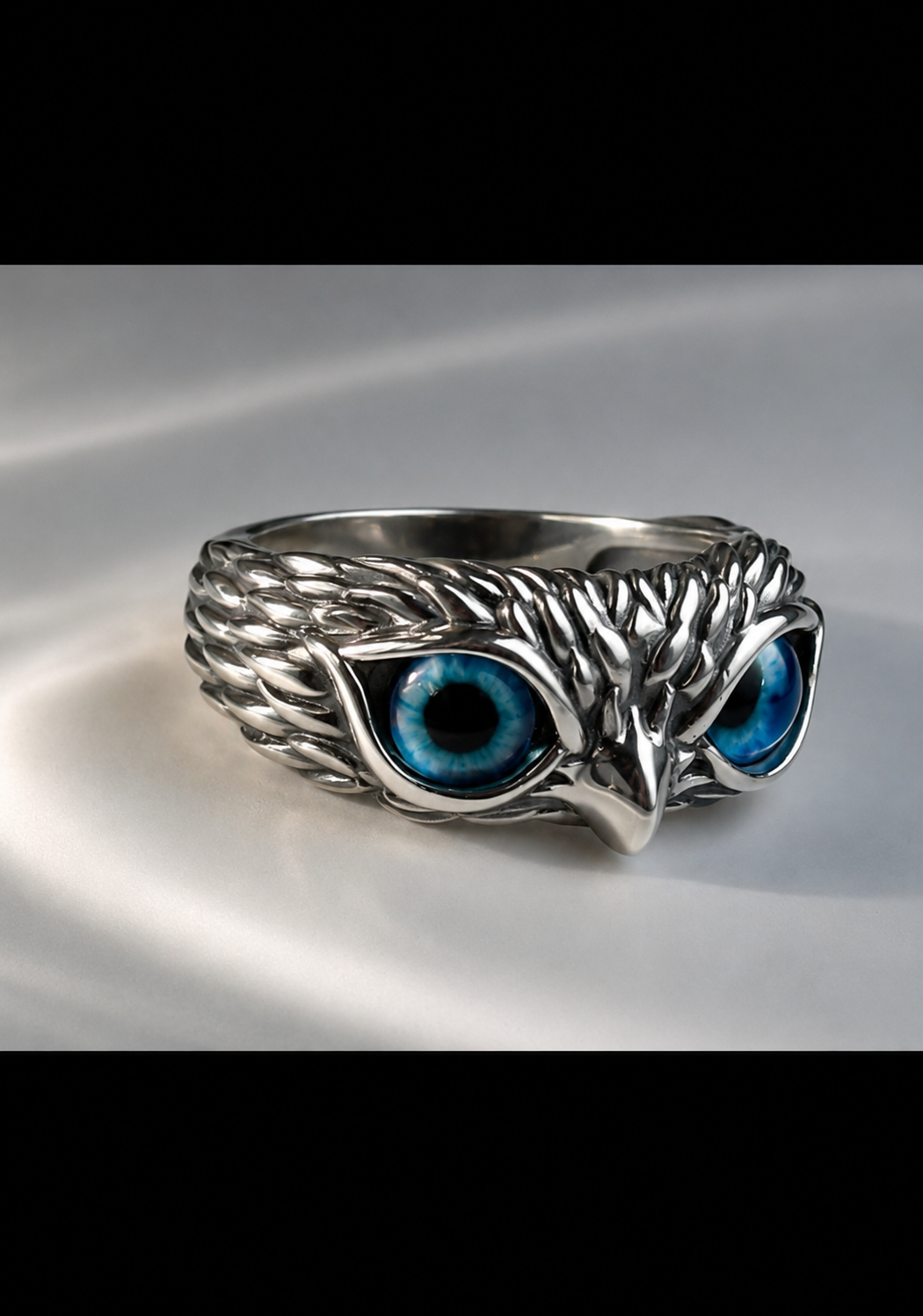 A silver owl head ring with detailed feather textures and vibrant blue enamel eyes, displayed on a light gray satin fabric.